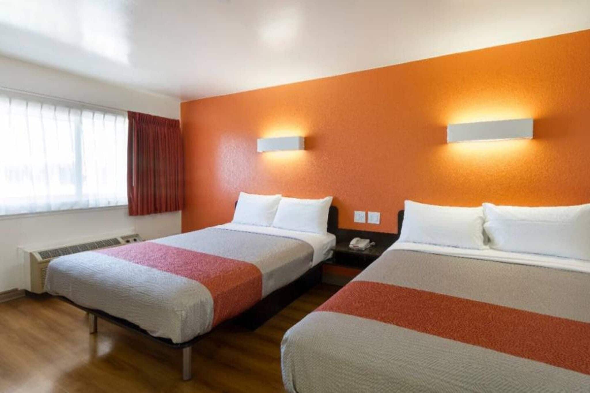 A hotel room with two beds, a window with curtains, a wall-mounted nightstand, and a thermostat.