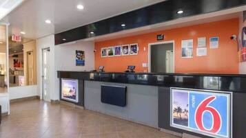 Een hotel receptie met een balie, een digitaal scherm en een muur met genummerde bordjes.