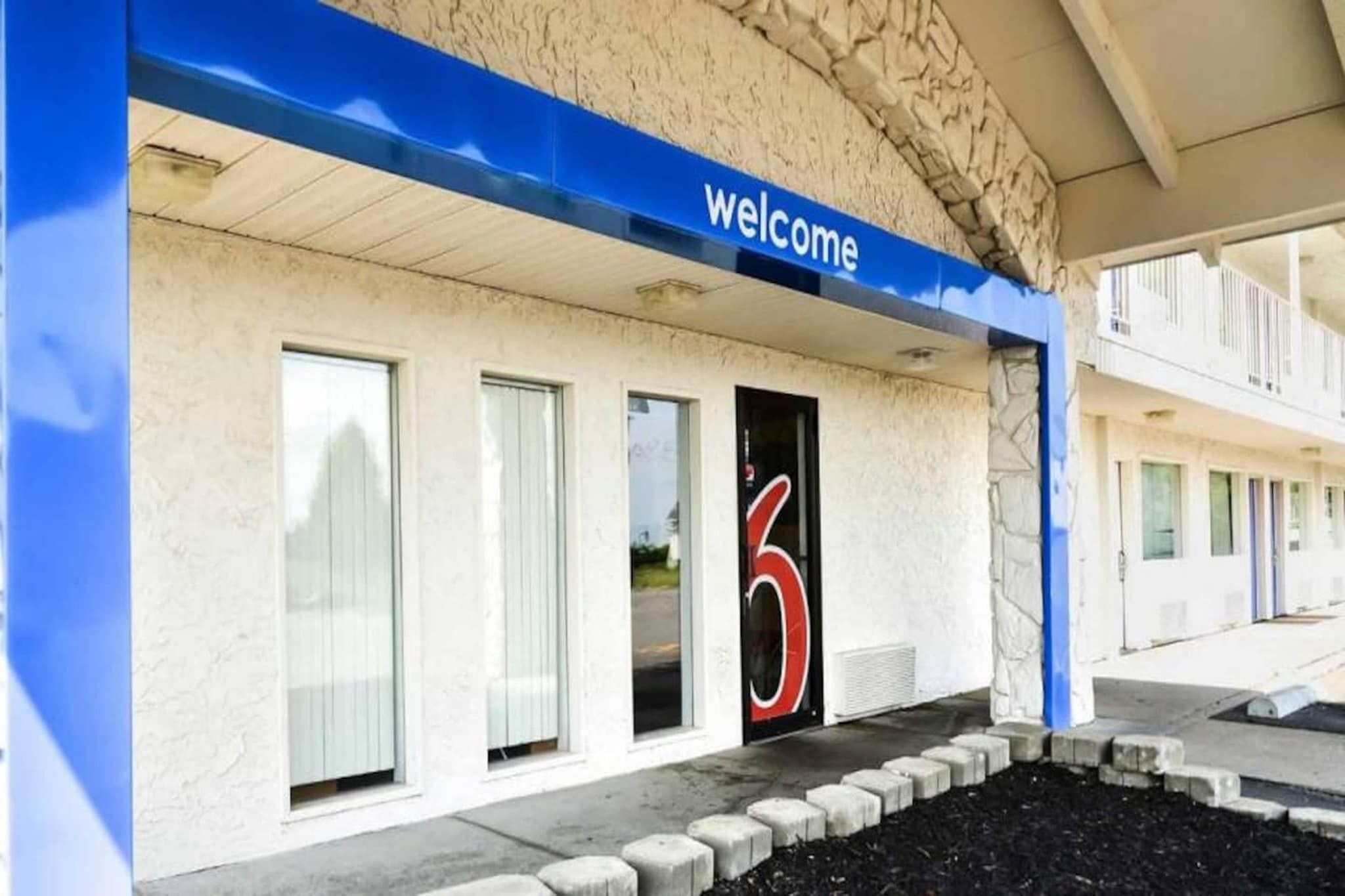 Entrada de um edifício com uma placa de 'welcome', o número '6' grande na porta e uma galeria coberta que leva a outros quartos.
