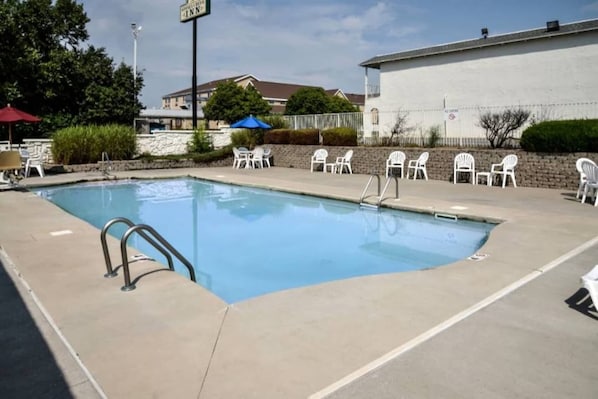 A rectangular swimming pool with a metal ladder on one side, surrounded by a concrete deck. There are white plastic chairs and a blue umbrella in the background. - Spacious room, 2 double beds | Accessible/Bathtub (Saint Joseph)
