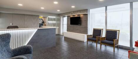 Eine Hotelhalle mit einer Rezeption, einer Sitzecke, großen Fenstern und einem an der Wand befestigten Flachbildfernseher.