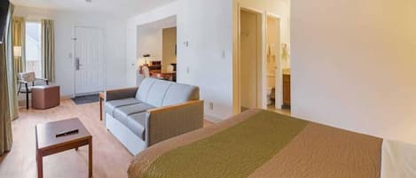 A hotel room with a bed, a sofa, a small table, and a chair. There is a door leading to a bathroom.