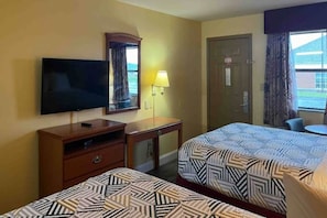 Quarto de hotel com duas camas, televisão, escrivaninha, luminária e porta.