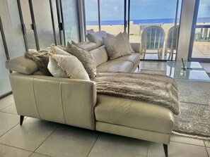 Un sofà de pell beix amb una manta de pell i coixins, ubicat en una habitació amb grans finestres que donen a una platja.