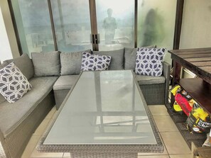 Een moderne woonkamer met een salontafel met glazen blad, een grijze bank met patroon kussens en een rieten bijzettafeltje met een zak chips.