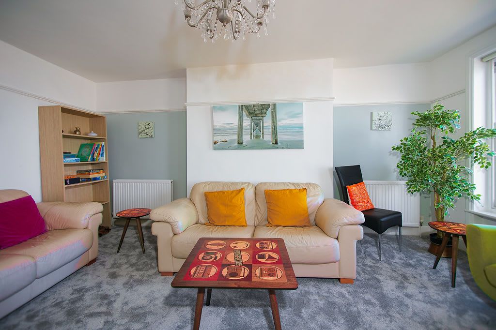 A living room with a beige sofa, a coffee table with a game, a bookshelf, a painting of a pier, and a potted plant.