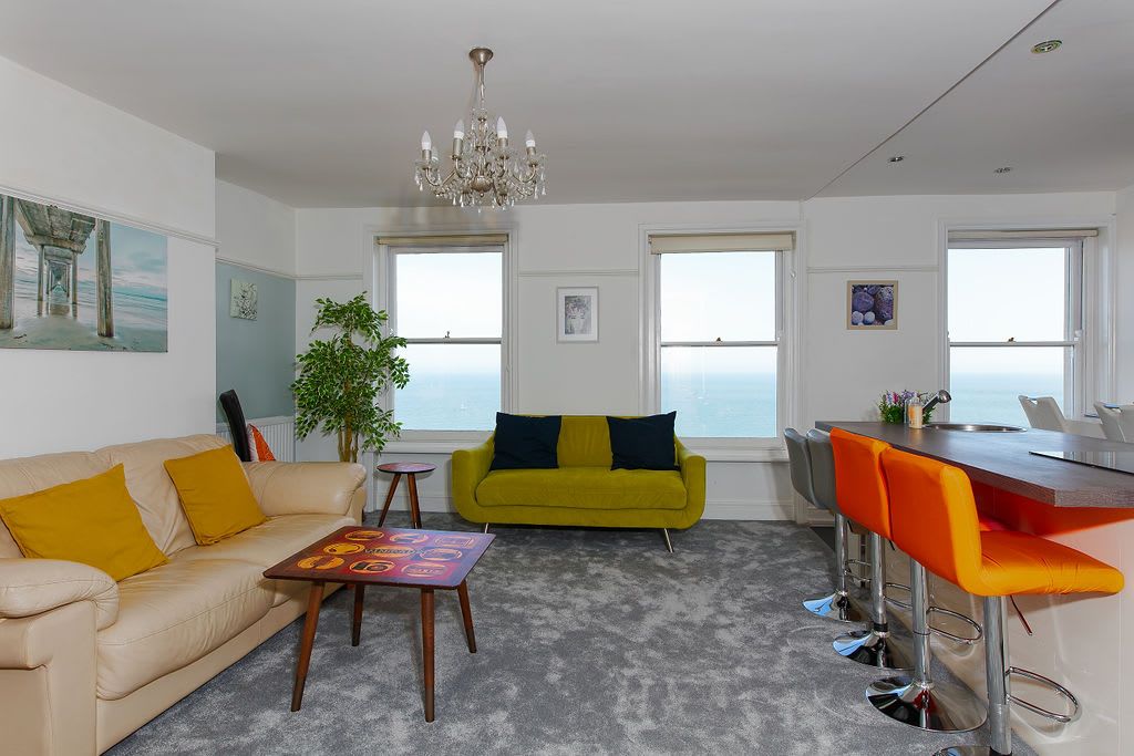 A modern living room with a sofa, a small table, a bar area with stools, and a view through large windows.