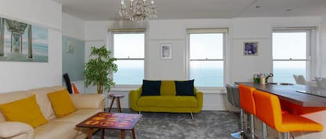 A modern living room with a sofa, a small table, a bar area with stools, and a view through large windows.