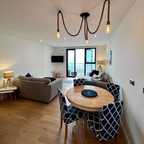 Ein modernes Wohnzimmer mit einer Couch, einem Esstisch mit Stühlen, einem an der Wand befestigten Fernseher und einem großen Fenster mit Blick.
