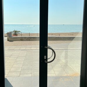 Blick durch eine Glastür mit Metallgriff auf eine gepflasterte Fläche, einen Sandstrand und das Meer im Hintergrund.