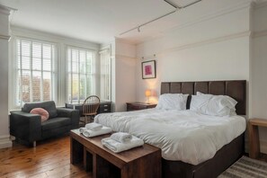 Een hotelkamer met een groot bed, een zithoek met een stoel en een tafel, en een raam met jaloezieën.