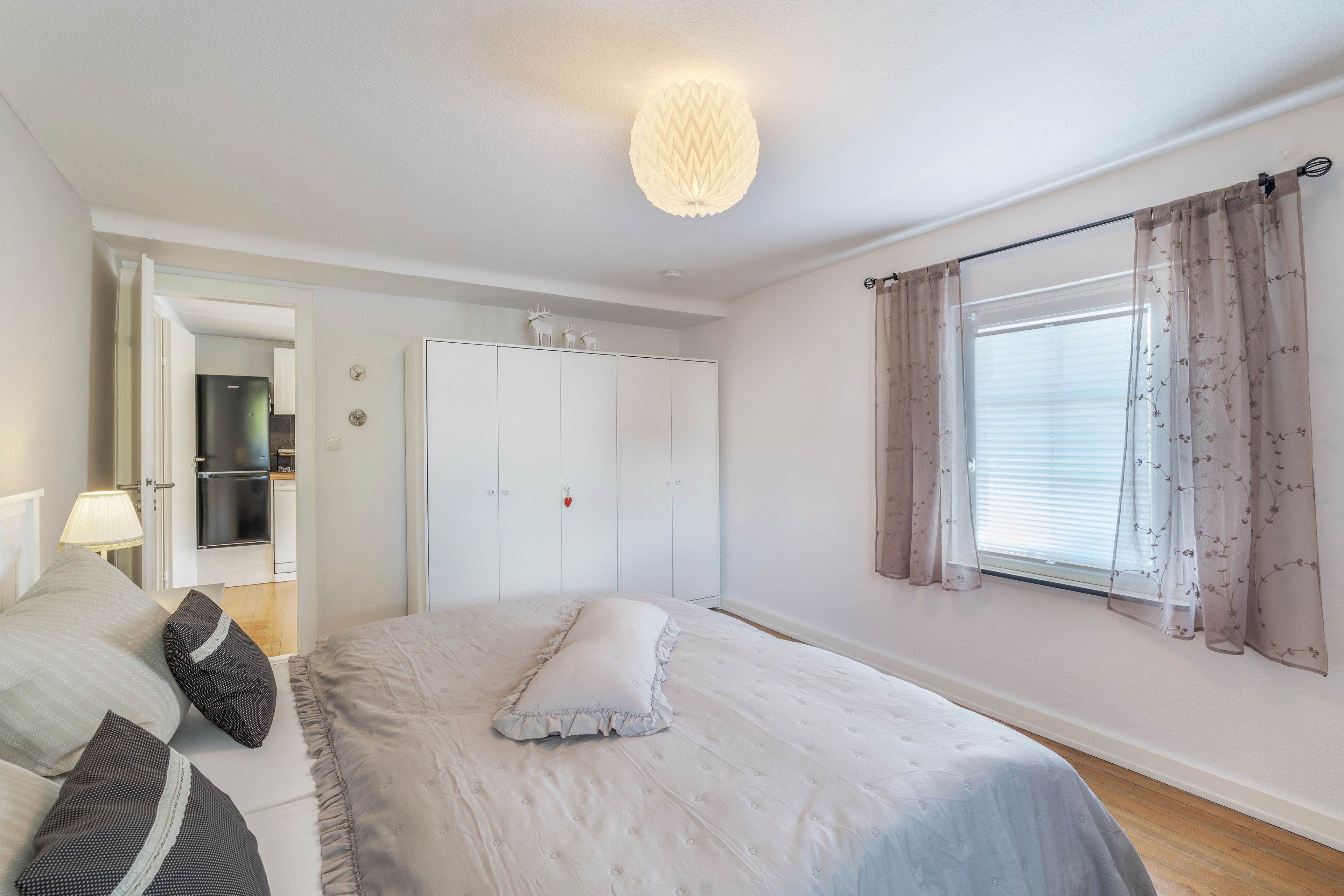 Ein Schlafzimmer mit Bett, Kleiderschrank, einem Fenster mit Vorhängen und einer Lampe.