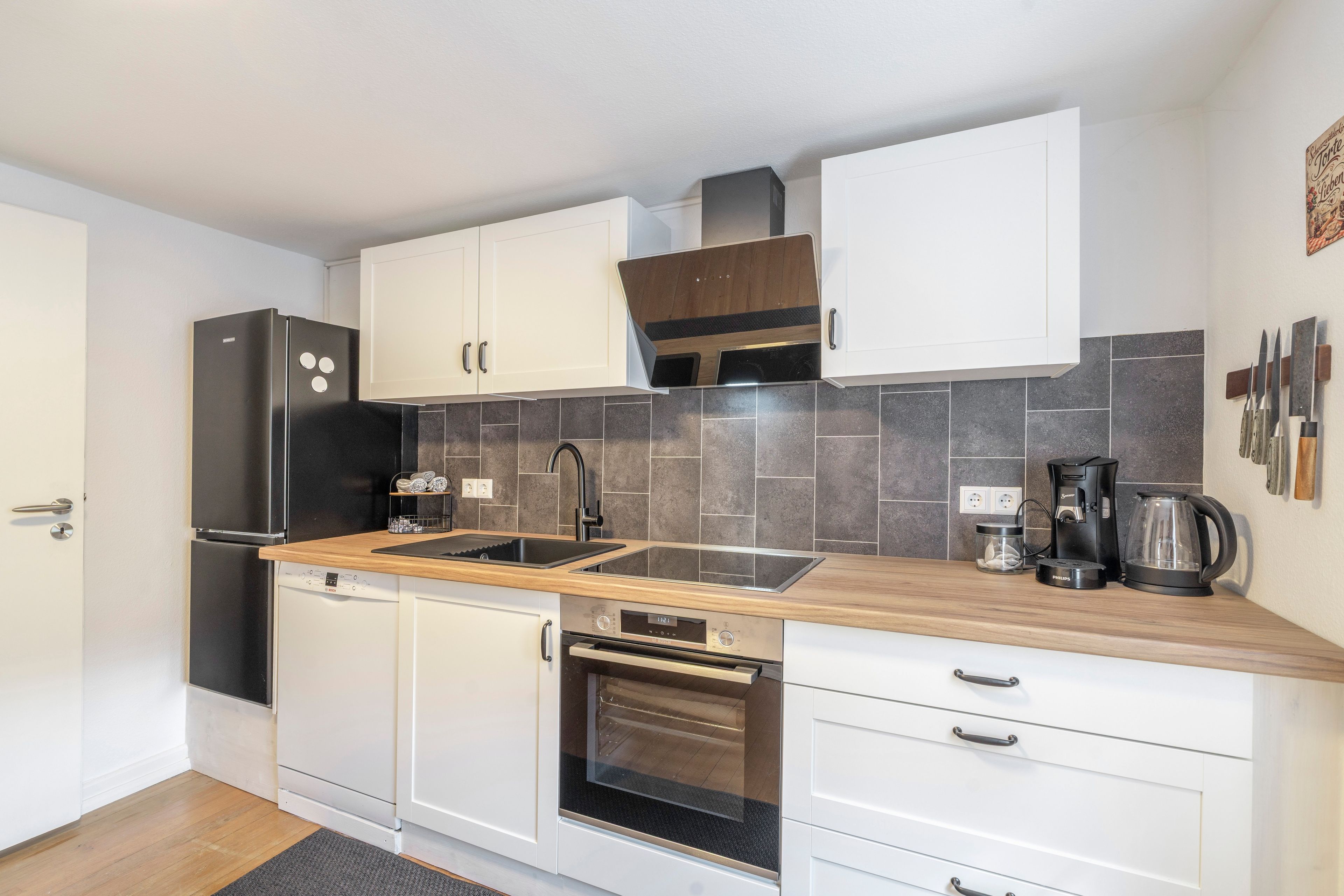 A compact kitchen with white cabinets, a black refrigerator, a built-in oven, a coffee maker, and a kettle.