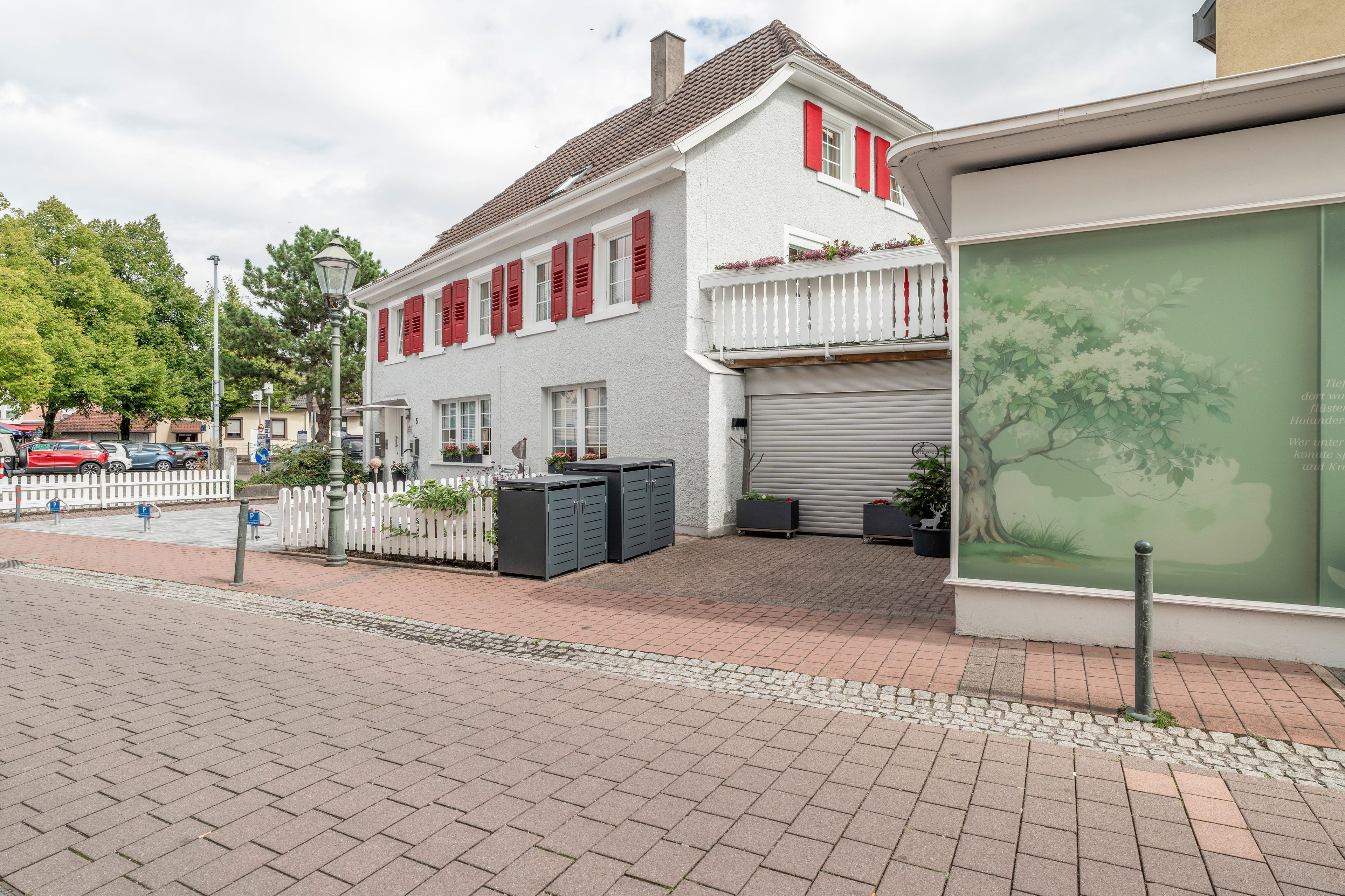 En tvåvåningsbyggnad med en vit fasad och röda fönsterluckor, en uppfart med grind och ett garage med stängd lucka.