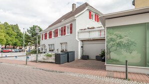 Un edificio de dos pisos con fachada blanca y contraventanas rojas, un camino de entrada con portón y un garaje con persiana cerrada.