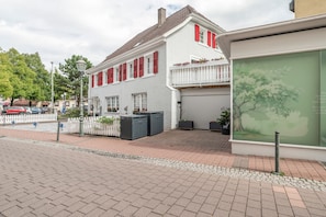 En tvåvåningsbyggnad med en vit fasad och röda fönsterluckor, en uppfart med grind och ett garage med stängd lucka.