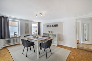 Un saló modern amb una taula de dinar, dues cadires, un radiador, una finestra amb cortines, una estanteta penjada a la paret amb objectes decoratius i una porta.