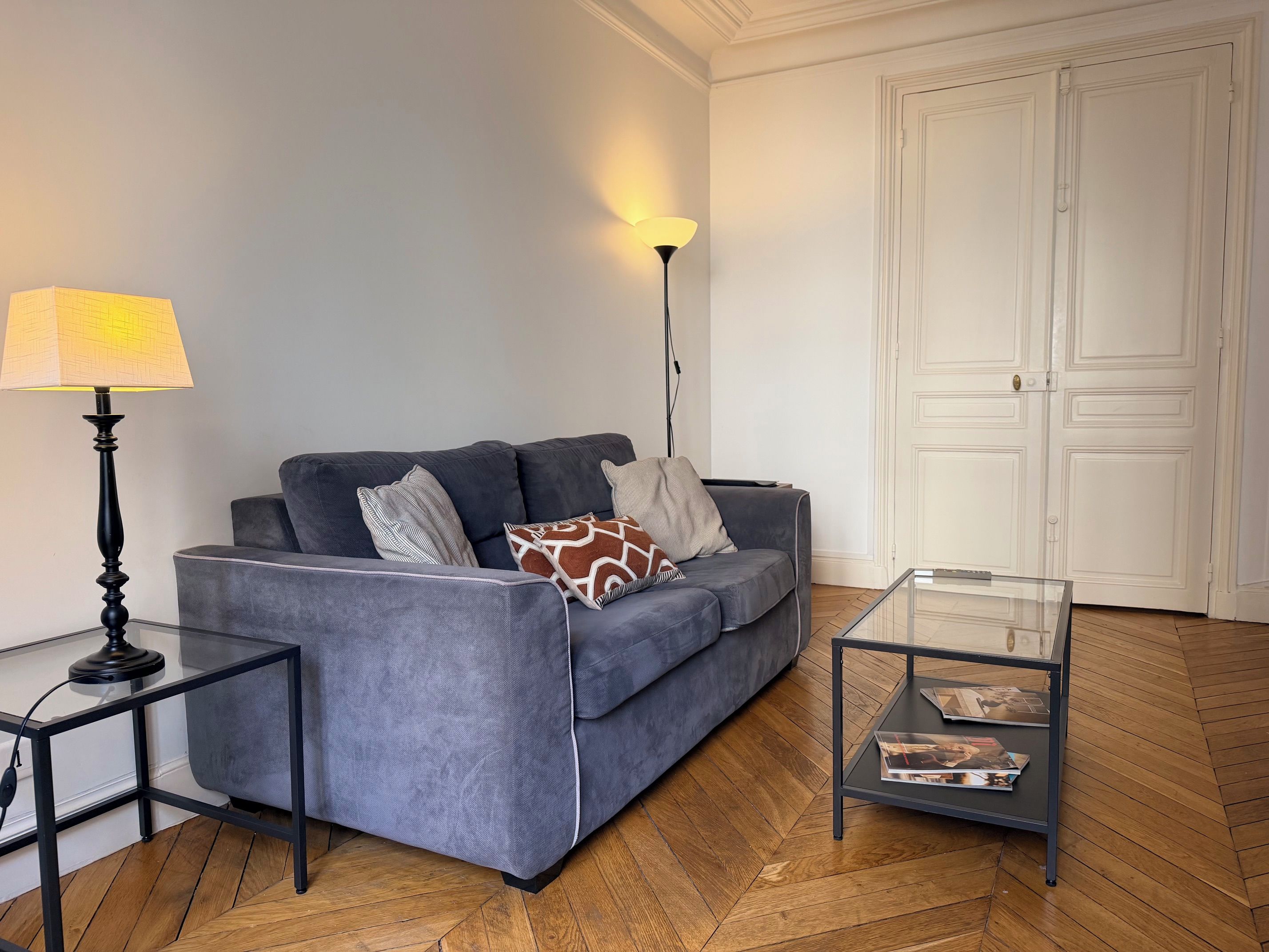 Een woonkamer met een grijze bank, een glazen salontafel, een vloerlamp en een witte deur.