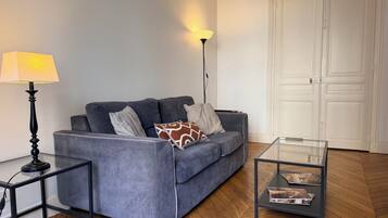 Een woonkamer met een grijze bank, een glazen salontafel, een vloerlamp en een witte deur.