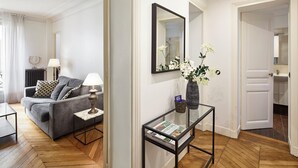 A modern living room with a grey sofa, a small side table with a lamp, and a glass coffee table. There's a mirror on the wall and a vase with white flowers on a side table. - Luxury 1BR steps away from Champs Elysees (Paris)
