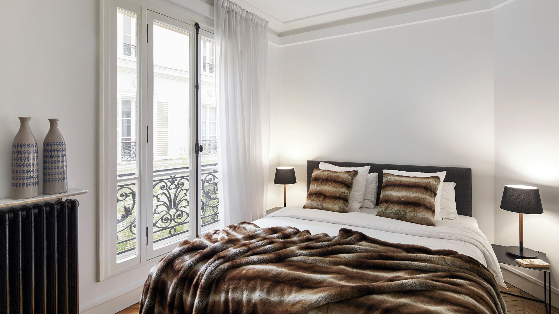 Een slaapkamer met een bed, twee bedlampen, een bont plaid en uitzicht buiten door een raam met gordijnen.