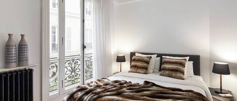 Une chambre à coucher avec un lit, deux lampes de chevet, un plaid en fausse fourrure et une vue sur l’extérieur par une fenêtre ornée de rideaux.