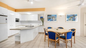 A modern kitchen with white cabinets, a central island, and blue chairs around a table. - Como Noosa 7 Noosaville (Noosaville)