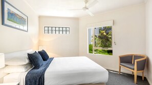 A hotel room with a bed, a chair, a window with a view of greenery, a ceiling fan, and a wall-mounted light fixture. - Como Noosa 7 Noosaville (Noosaville)