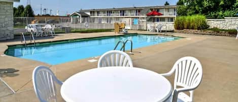A swimming pool with a patio area, including a table and chairs, and a building with a balcony in the background.