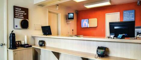 Un espace d’accueil d’hôtel comprenant un comptoir, une machine à café, un téléphone et un écran de télévision.