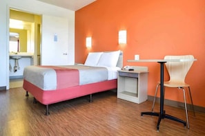 Habitación de hotel con cama, mesita de noche, silla y un baño visible a través de una puerta abierta.