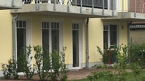 A two-story building with a flat roof, multiple windows, and a small garden area with young trees. - Comfortable "Lachmöwe" vacation apartment, 62 square meters, with a large terrace featuring a beach chair and a parking space (Börgerende-Rethwisch)