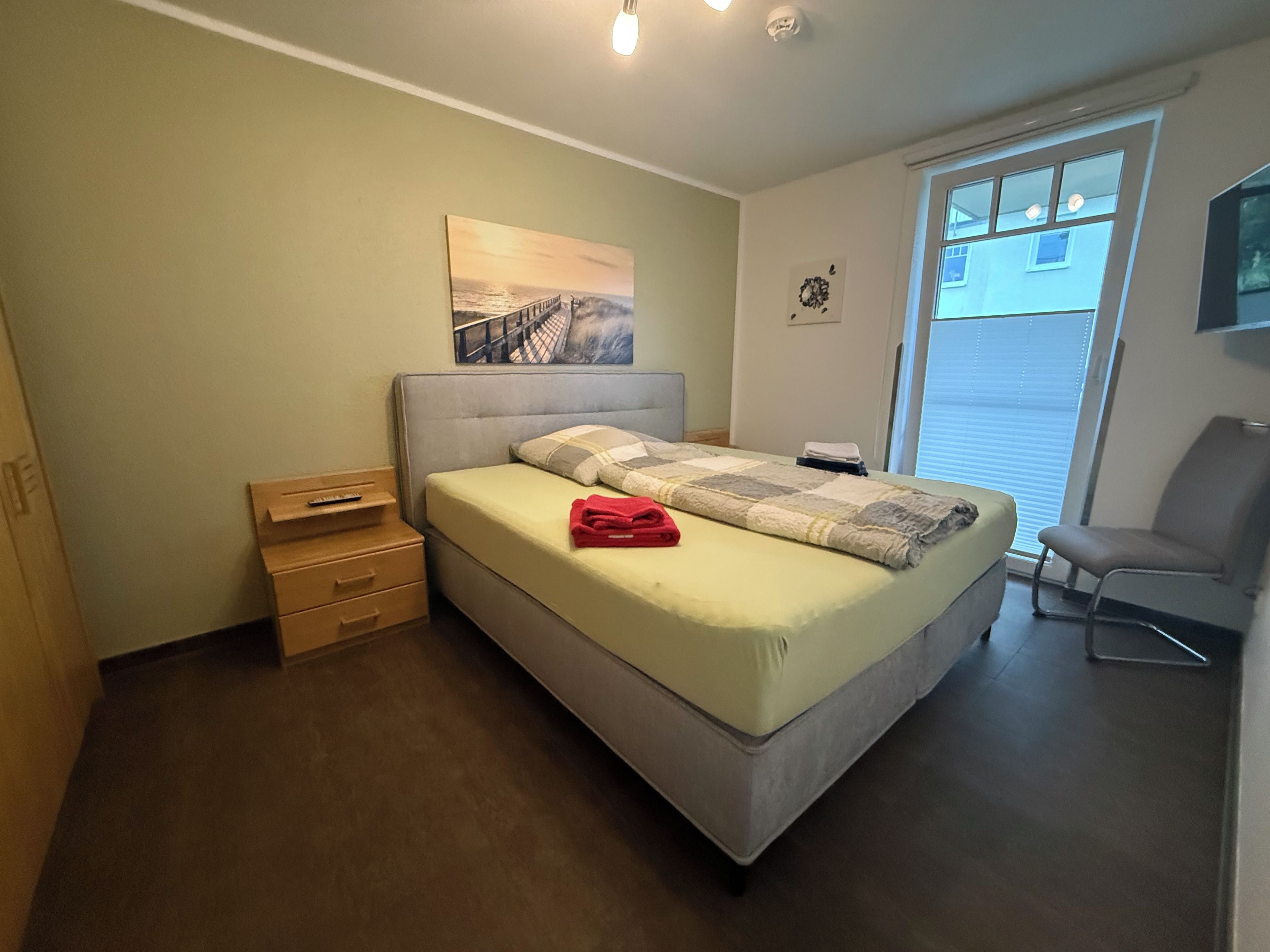 A bedroom with a bed, a nightstand, a chair, a window, and a painting on the wall.
