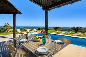 A dining table set for two with a view of the sea, a pool, and outdoor seating. - Villa Aqua in Lachania (Rhodes)