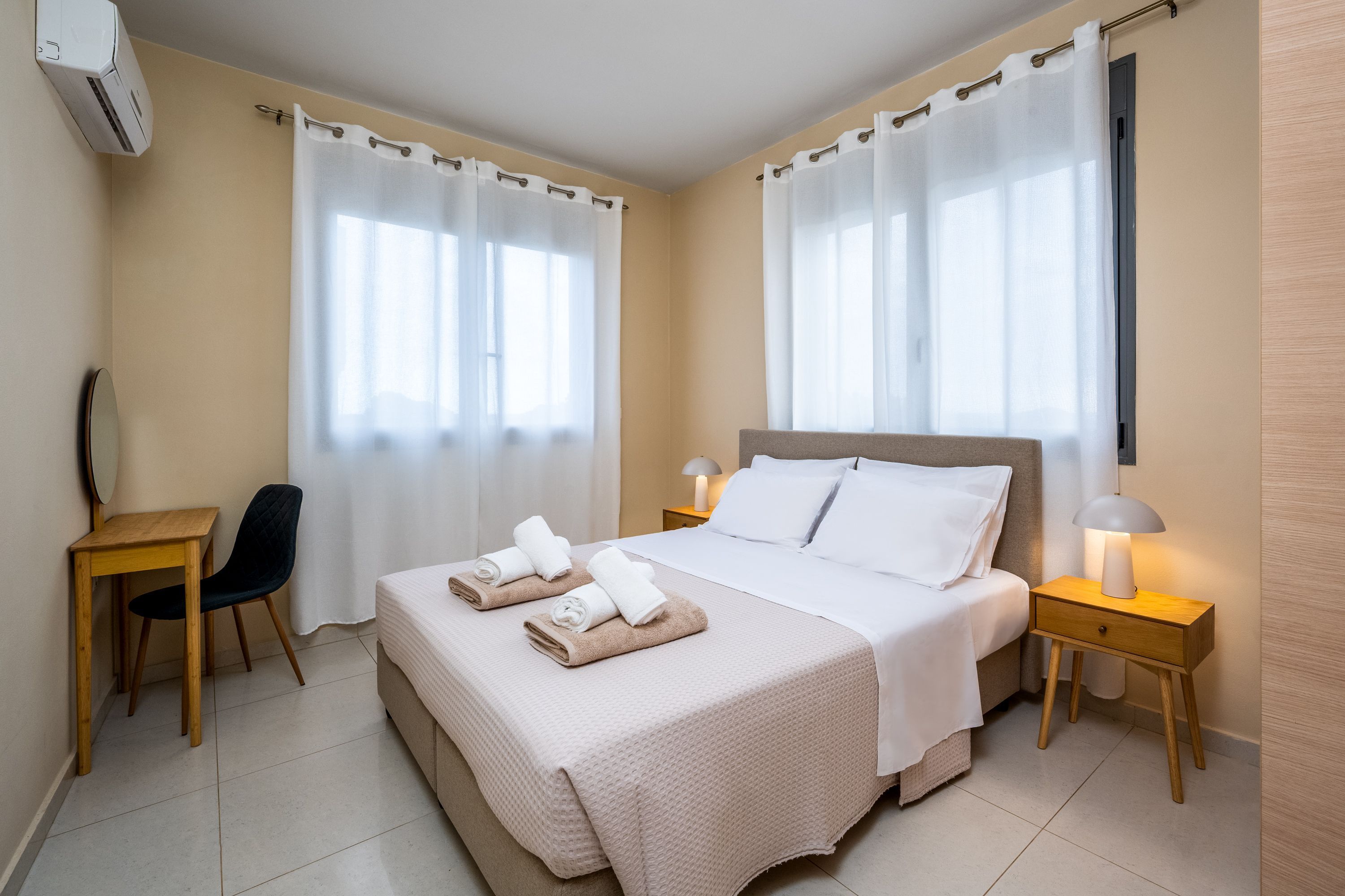 A hotel room with a bed, two bedside tables, a desk, a chair, and a window with curtains.