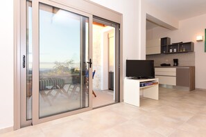 A modern living space with a sliding glass door leading to a balcony, a flat-screen TV on a stand, and a kitchenette area. - Mon Emvasia Margarites Villa B (Mylopotamos)