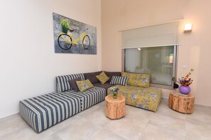 A modern living room with a sectional sofa, a painting of a bicycle, and a window with blinds. - Mon Emvasia Margarites Villa B (Mylopotamos)