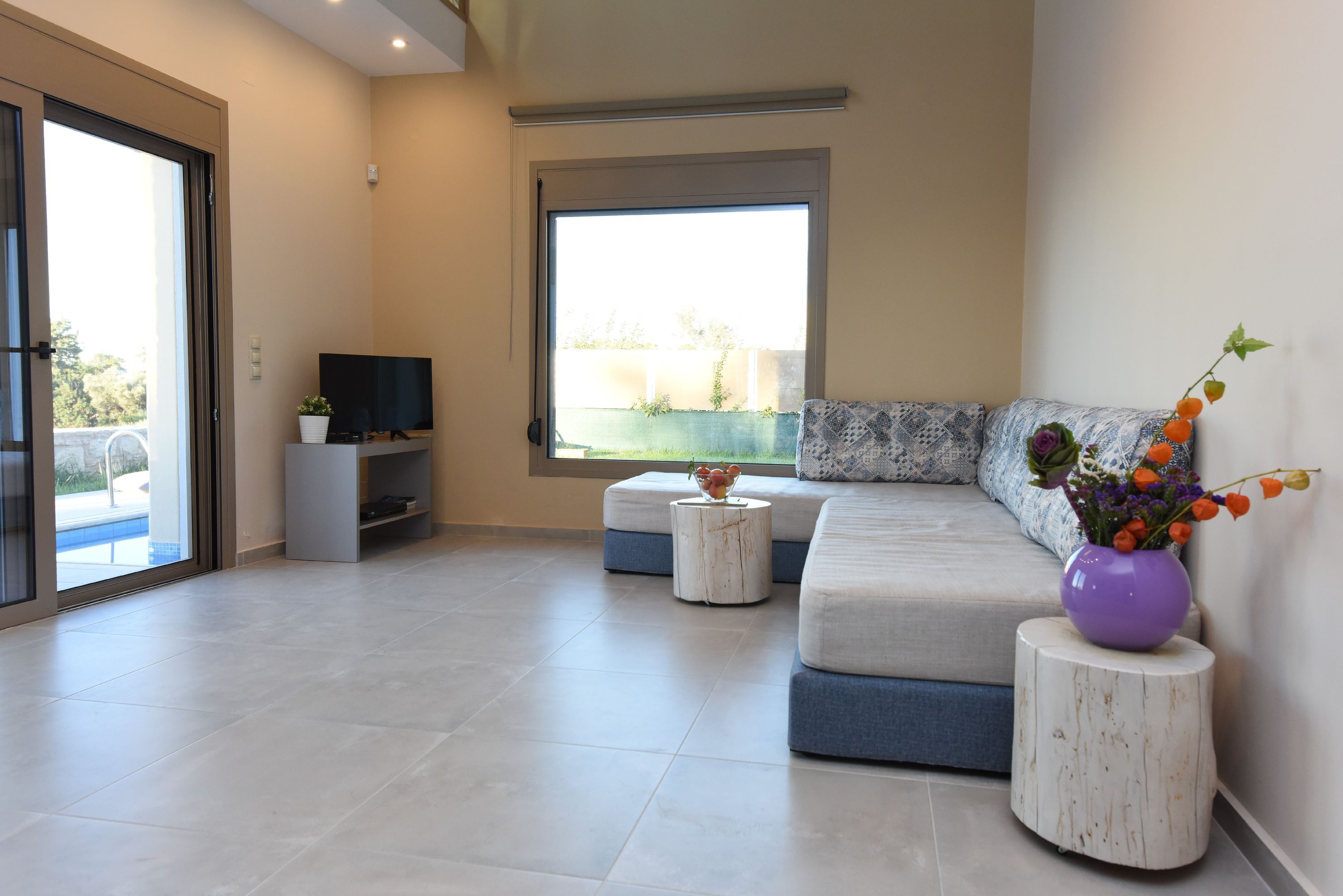 A modern living room with a large window, a flat-screen TV, a sofa, and a small side table with a vase of flowers.