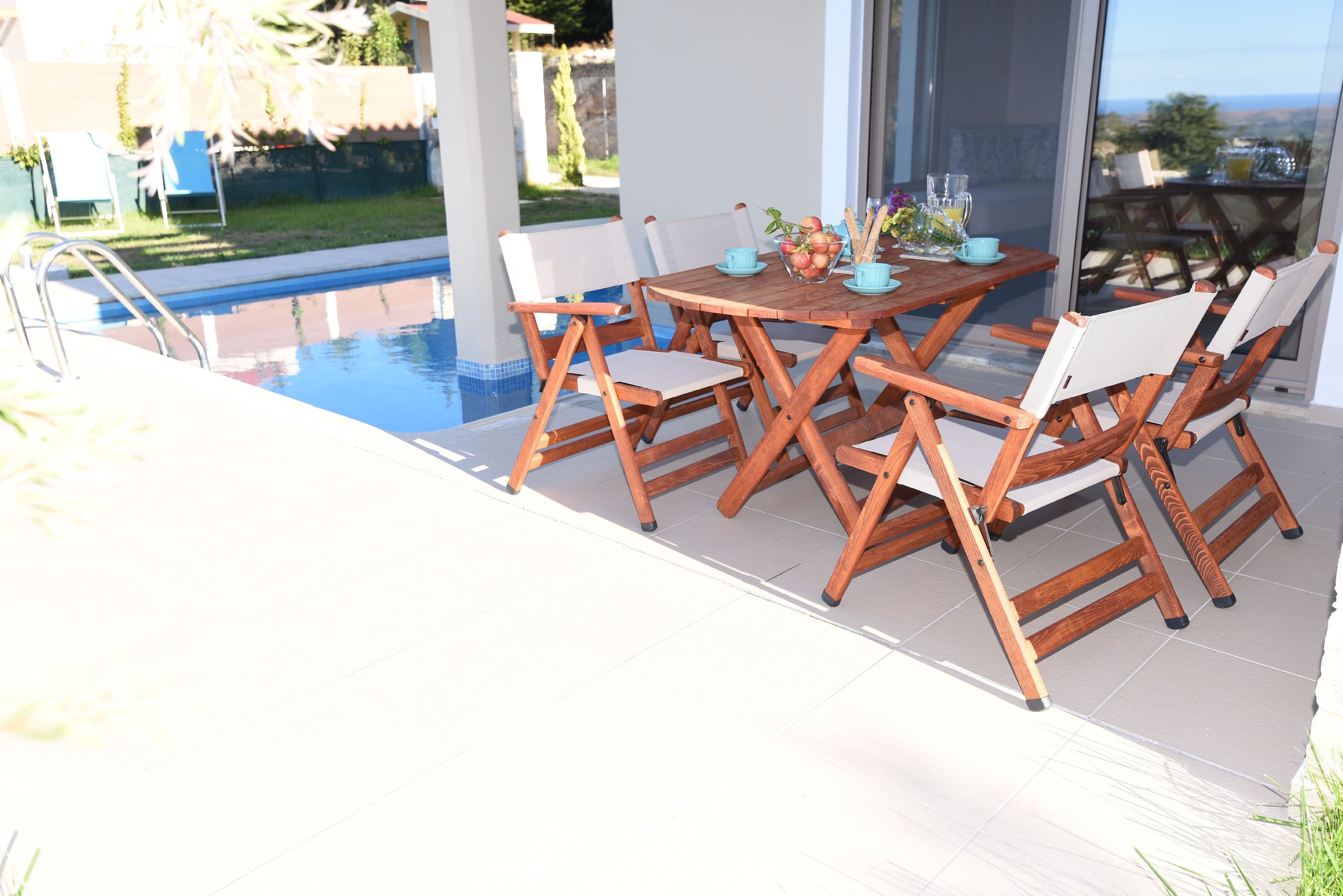 Un área de comedor exterior con piscina, sillas de madera y una mesa dispuesta con tazas azules y un bol de fruta.