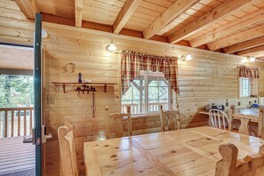 Interior de una cabaña de madera con una mesa de comedor, sillas, una ventana con cortinas y una pequeña cocina.