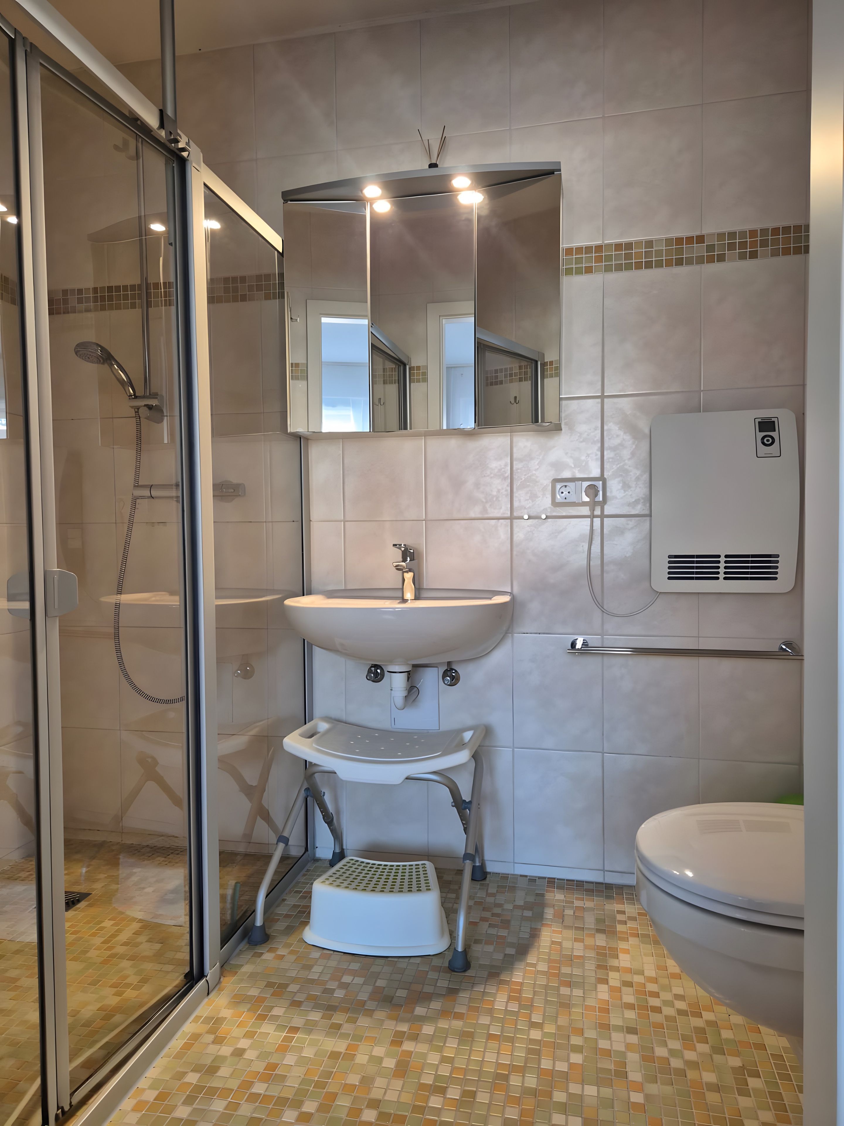 Ein Badezimmer mit Duschkabine aus Glas, einem Waschbecken mit Spiegel darüber, einer Toilette und einem kleinen Trittschemel.