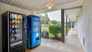 Ein Getränkeautomat und ein digitales Display in einem überdachten Außenbereich mit Betonboden und Blick auf den Parkplatz.