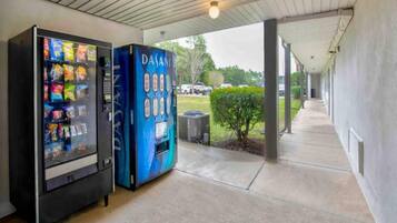 A vending machine and a digital display in a covered outdoor area with a concrete floor and a view of the parking lot.