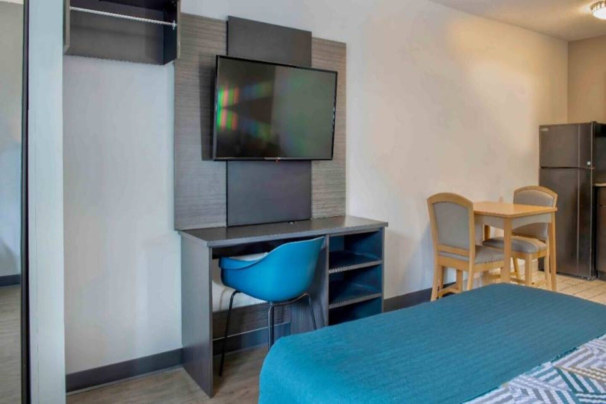 A hotel room with a bed, a desk with a chair, a television mounted on the wall, and a refrigerator.