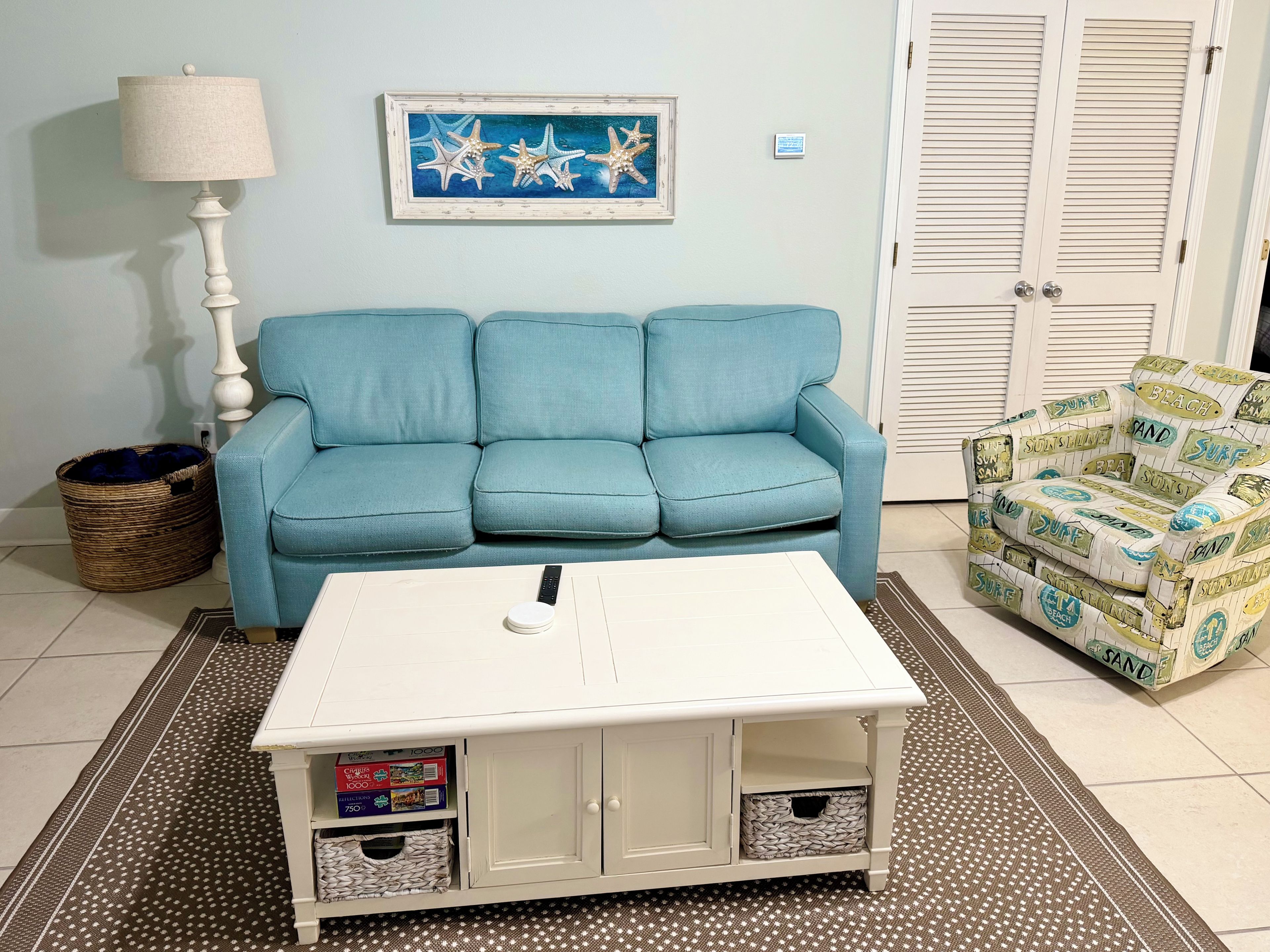 A living room with a blue sofa, a white coffee table, a wicker basket, and a beach-themed armchair.