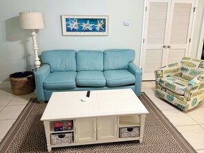 A living room with a blue sofa, a white coffee table, a wicker basket, and a beach-themed armchair. - 3-bedroom pet-friendly condo in Orange Beach, sleeps 8 (Orange Beach)