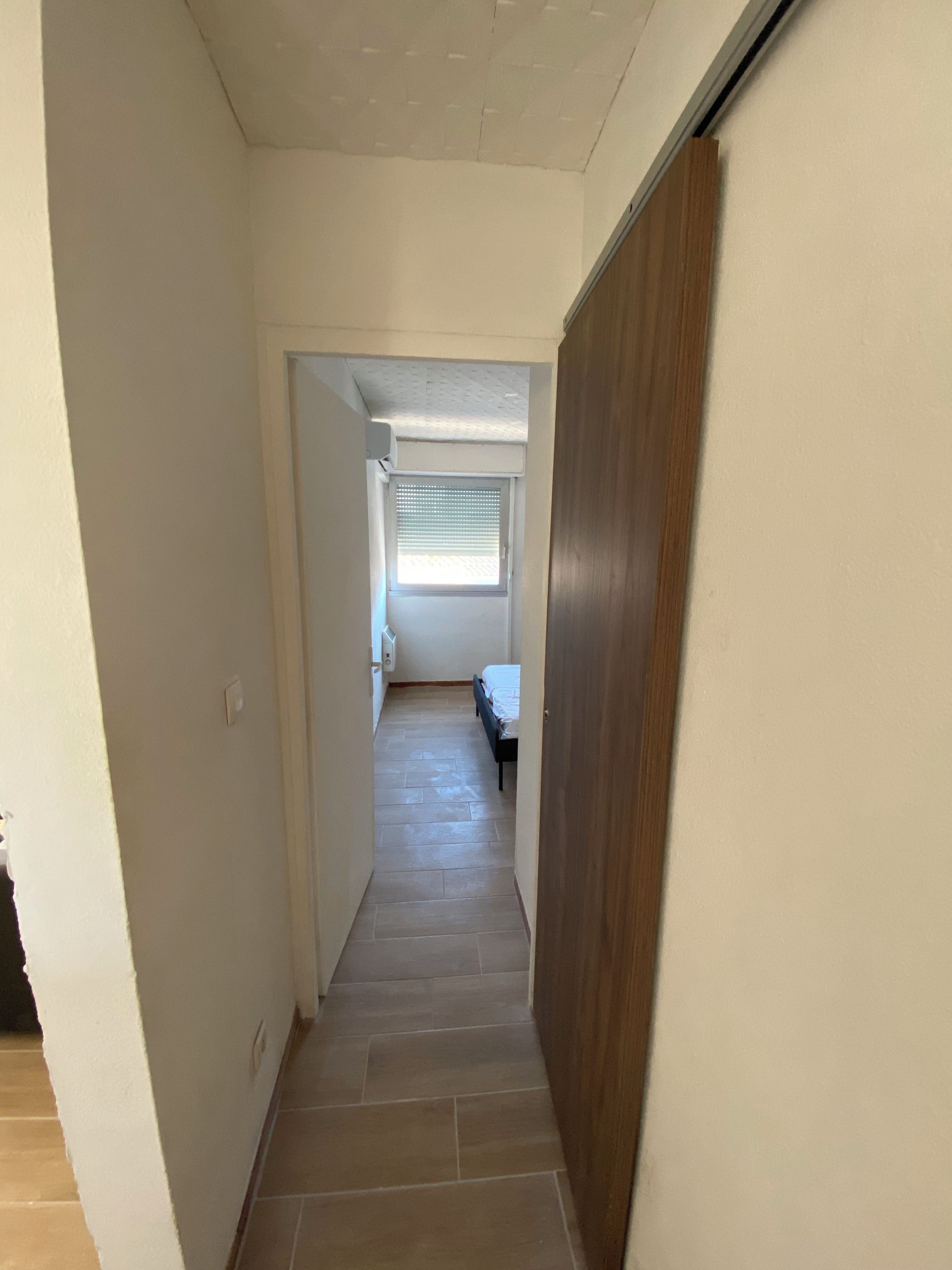 Un couloir étroit, avec une porte en bois sur la droite, une fenêtre avec des stores au fond, et un lit visible au loin.