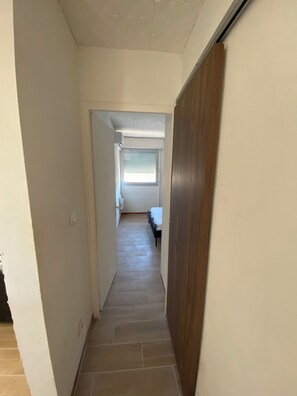 A narrow hallway with a wooden door on the right, a window with blinds at the end, and a bed in the distance. - Panoramic Sea View – Cozy Renovated Apartment (Hérault)