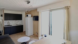 Een compacte keuken met een koelkast, magnetron en een spoelbak met een marmeren aanrecht. Er is een eethoek met een tafel en stoelen, en een groot raam met gordijnen.