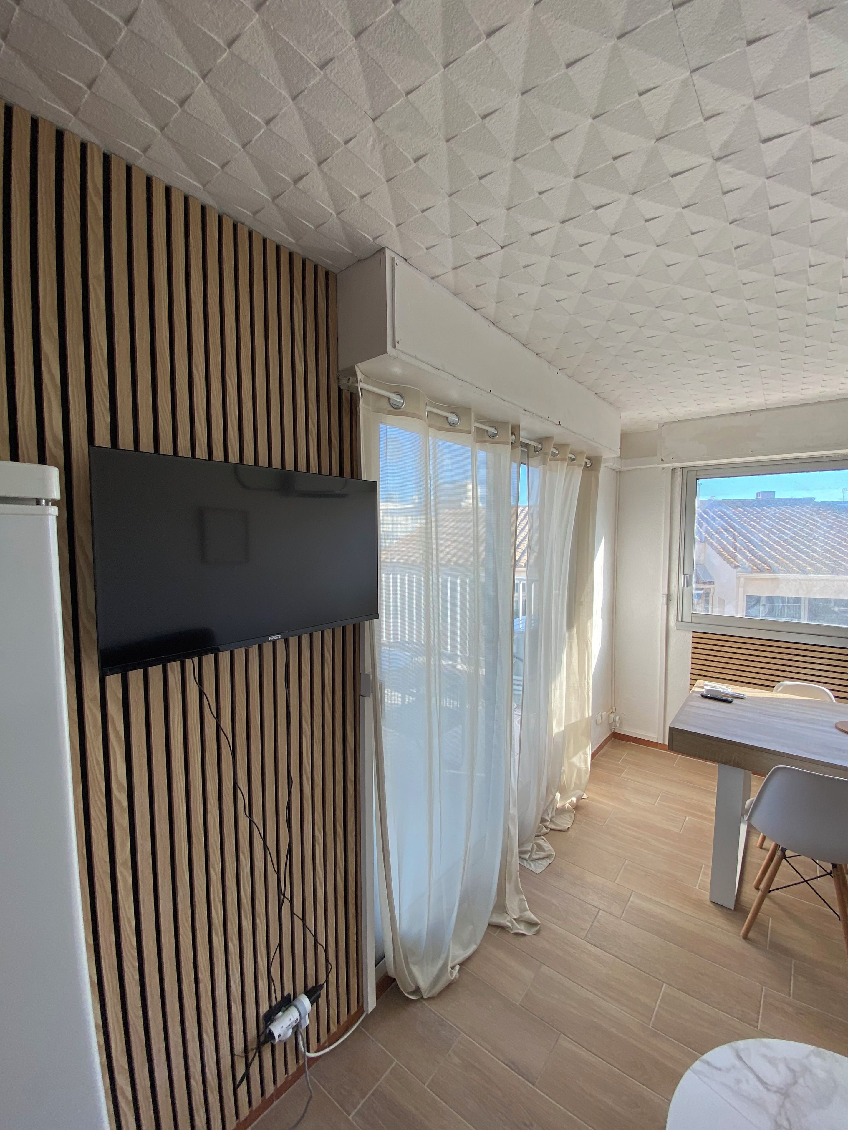 A modern living room with a flat-screen TV mounted on a wooden panel wall, a dining area with a table and chairs, and a large window with curtains.